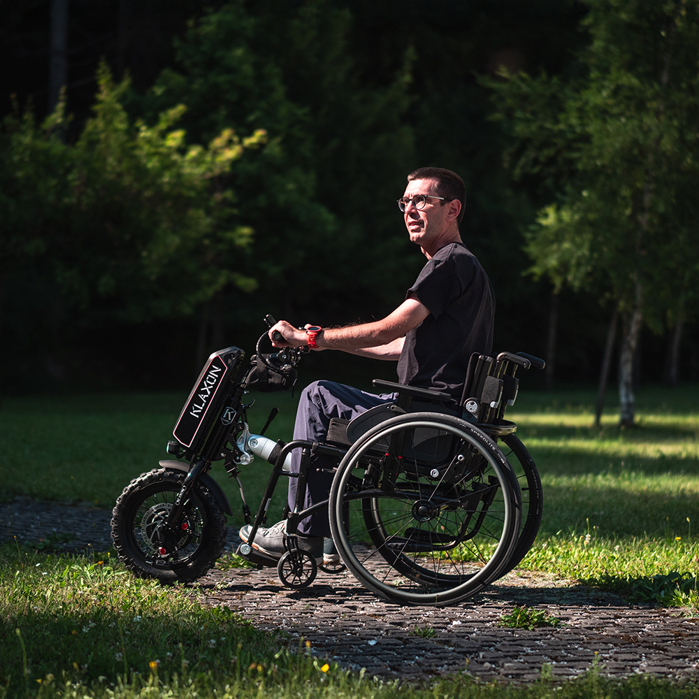 A user in a manual wheelchair utilizing the Klaxon Klick Power electric handbike attachment in an outdoor lifestyle setting, highlighting its 750W motor and 14-inch wheel for smooth, versatile mobility on paved and light off-road paths.