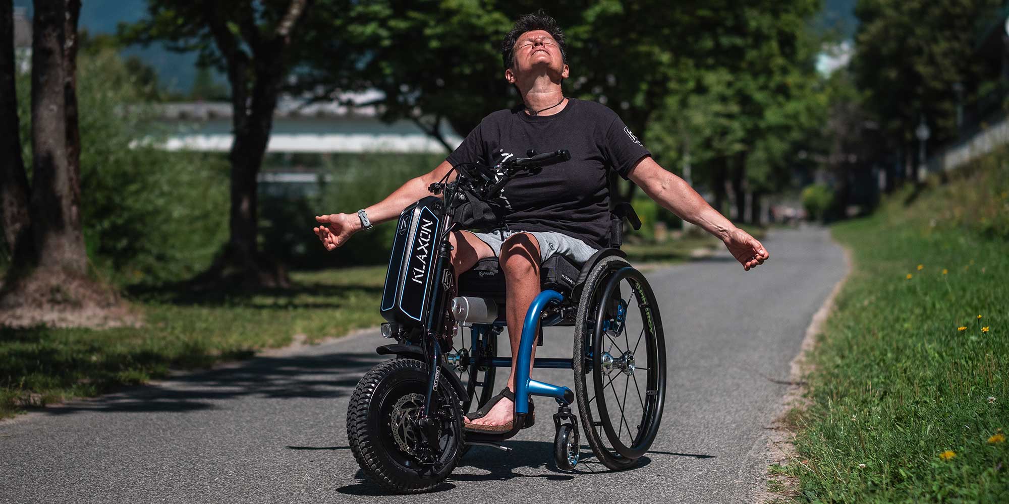 Alt Text: A wide lifestyle banner showing a user in a manual wheelchair with the Klaxon Klick Scrambler electric handbike attachment, demonstrating its rugged off-road performance and power on an outdoor gravel path.