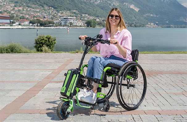 woman riding a Rehasense PAWS ECO Travel LX wheelchair power add-on along a waterfront promenade.