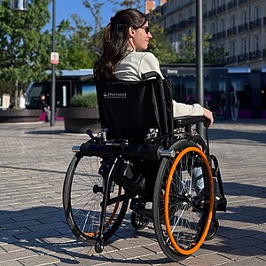 Gemini said In this sunlit urban scene, a woman with long dark hair and sunglasses sits in a Motion Composites manual wheelchair, seen from a rear three-quarter perspective as she looks across a paved city plaza. The wheelchair is outfitted with a Benoit Systemes Light Drive 3 power add-on, a compact black motor unit mounted behind the backrest that uses rollers to power the rear wheels. The chair itself features a sleek black frame accented by vibrant bright orange handrims on the large rear wheels. In the background, a modern tram and city buildings are visible under a clear blue sky, highlighting the system's use for outdoor mobility.