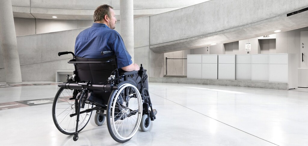 Landscape banner of a man in a manual wheelchair using the Benoit Systèmes Light Drive 3 XL power add-on in a modern, spacious building lobby.
