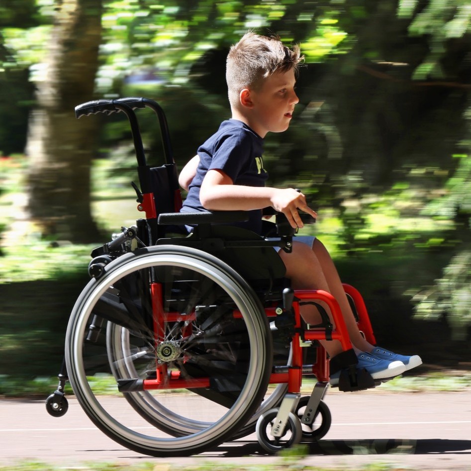 Side view of a young boy quickly navigating an outdoor path using the Light Drive Mini 3 wheelchair power add-on.