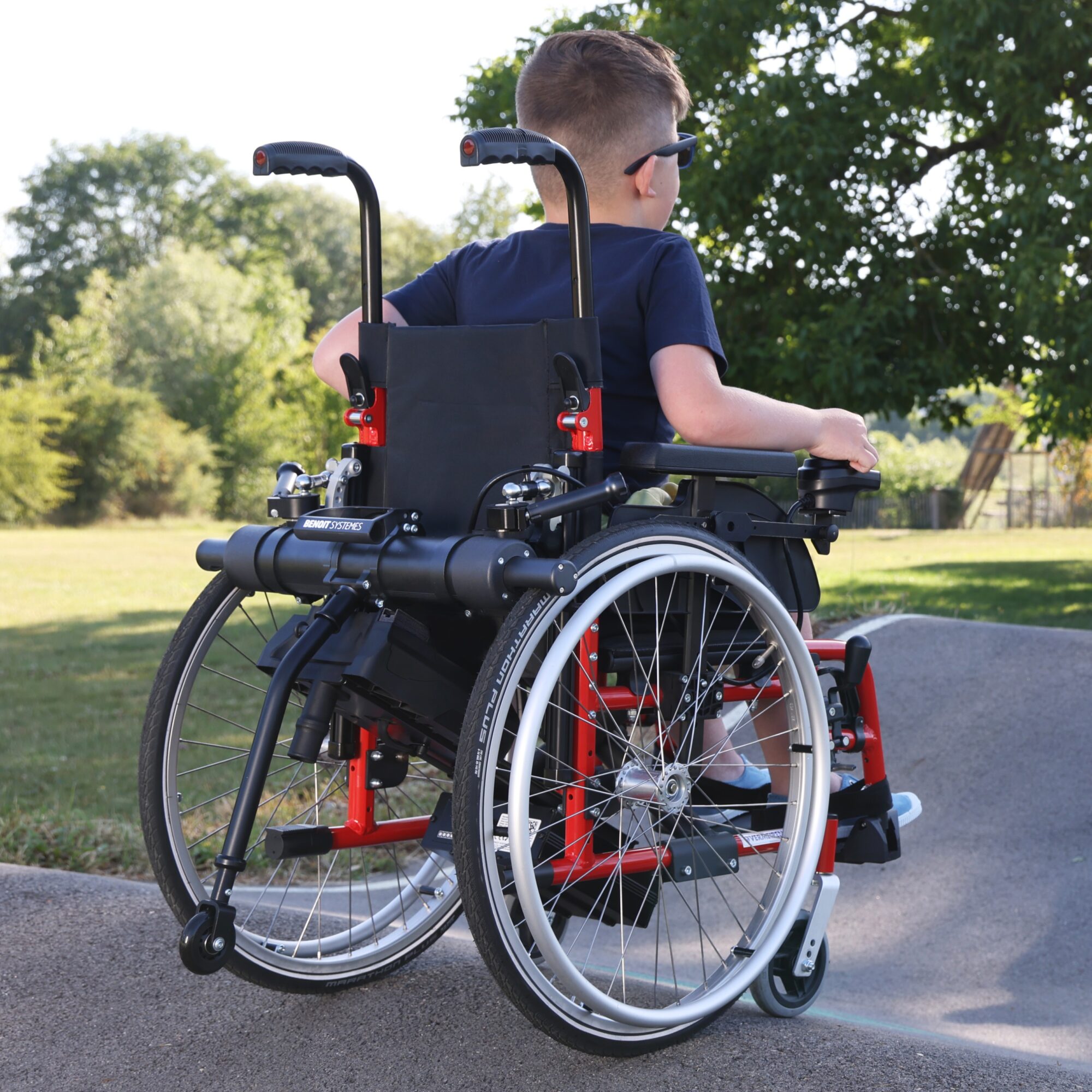 Child using a red manual wheelchair equipped with a Benoit Systèmes Light Drive Mini 3 power add-on at an outdoor park.