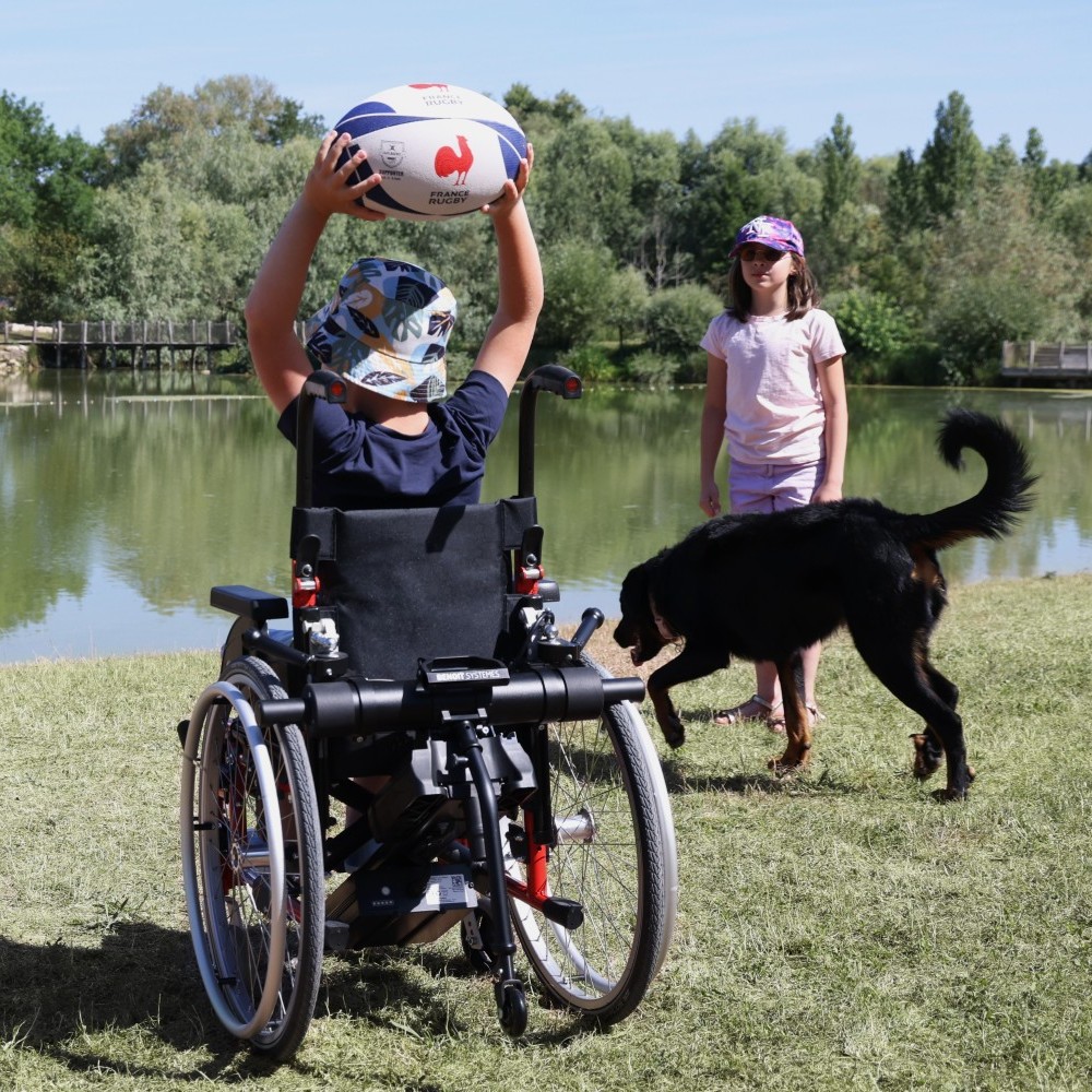 A child in a manual wheelchair with a Benoit Systèmes Light Drive Mini 3 power add-on plays with a rugby ball by a lake.