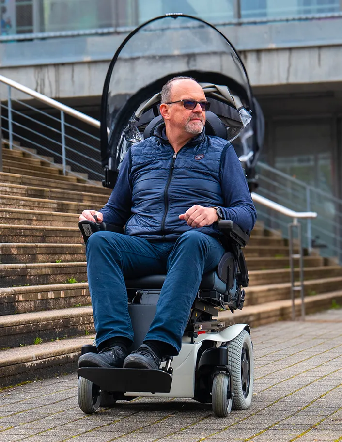 A man wearing a blue quilted vest and sunglasses sits in a power wheelchair with the Veltop Cosy 3 canopy retracted. The transparent panels and fabric are folded back behind the headrest, showing how the system allows the user to enjoy an open-air experience and clear visibility when protection is not needed.