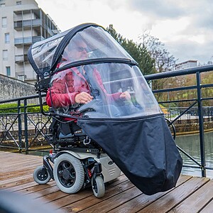 A close-up, eye-level shot of a woman in a red jacket using a power wheelchair on a wet wooden bridge. The Veltop Cosy 3 is fully deployed, and visible raindrops cling to the transparent windshield. The man is completely shielded from the rain by the overhead canopy, front windows, and black leg apron.